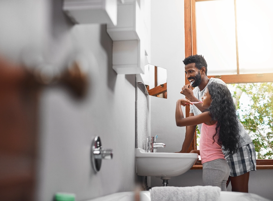 A father and daughter wash their teeth together in a bathroom with a large window