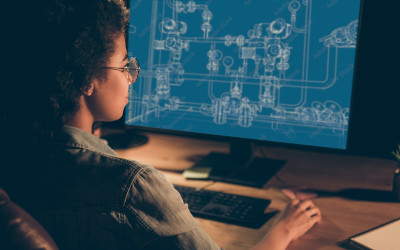 A woman sites at a computer with the blueprints for a series of pipes on the screen