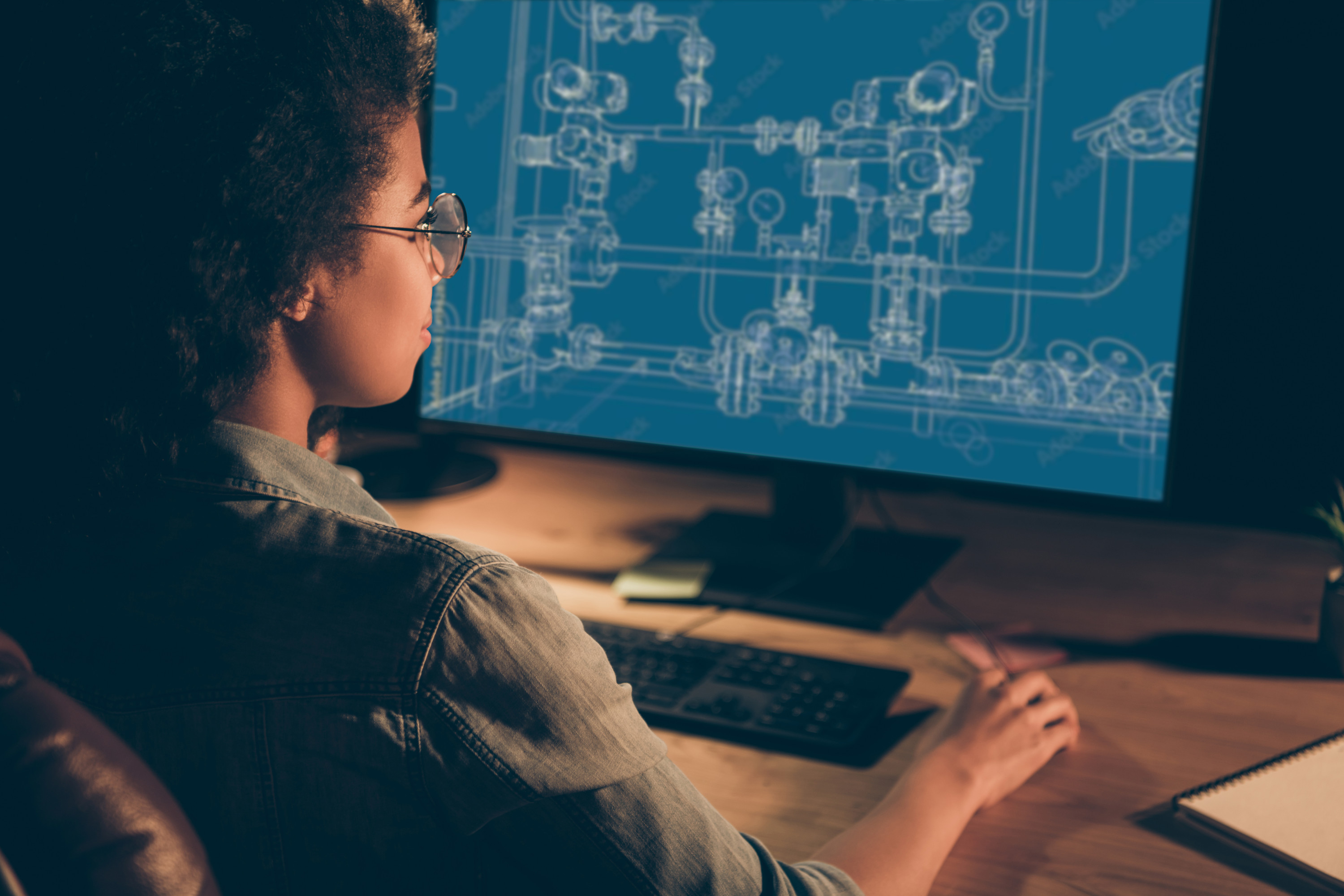 A woman sites at a computer with the blueprints for a series of pipes on the screen