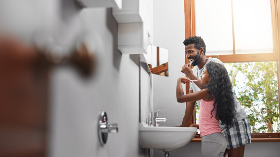 A father and daughter wash their teeth together in a bathroom with a large window